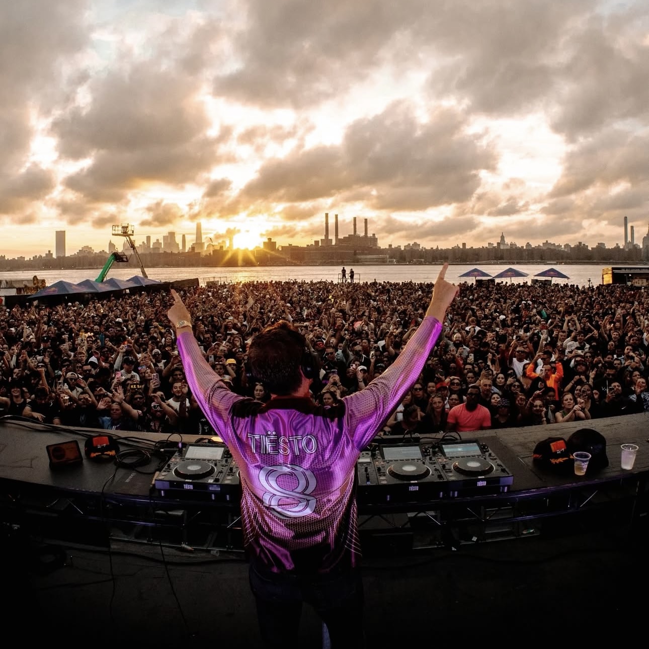 DJ Tiesto performing at outdoor waterfront festival with massive crowd at sunset