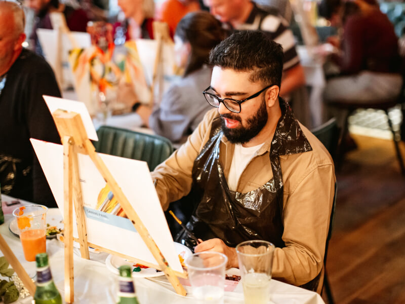 Person engaged in a painting workshop, focused and enjoying the creative process in a social setting with other workshop participants in the background